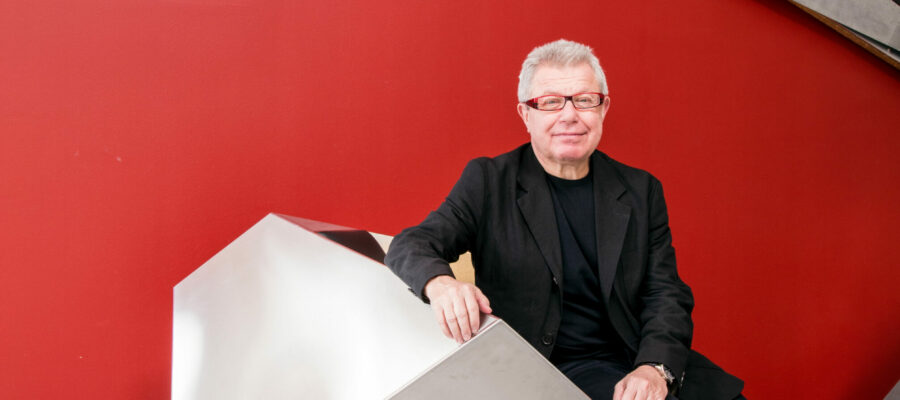 Foto von Daniel Libeskind – Aufnahme vor rotem Hintergrund, sitzend auf einer geometrischen Form