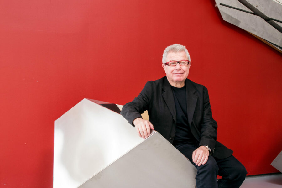 Foto von Daniel Libeskind – Aufnahme vor rotem Hintergrund, sitzend auf einer geometrischen Form